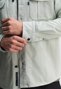 Lichtgrijze button-up shirt, met een borstzak, knopen bij de manchetten en een zijkraaglabel. Glad materiaal met een ontspannen pasvorm en naaddetails.