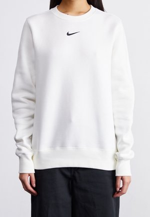 Persoon draagt een effen witte sweatshirt met lange mouwen en een klein zwart Nike-logo, geplaatst in het midden onder de halslijn.
