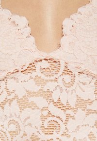 Krajkový BODY v jemně růžové barvě s hlubokým V-výstřihem. Obsahuje složité květinové vzory a zvlněné okraje, což vytváří jemnou strukturu.