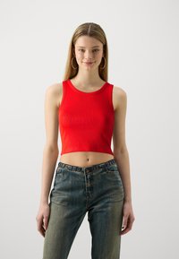 Rød ribbestrikket crop-top med vid halsringning og uten ermer, sammen med falmede blå jeans med knappelukking i livet. Enfarget bakgrunn.