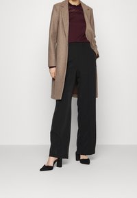 Lungo cappotto beige su una maglia bordeaux con dettagli in pizzo, abbinato a pantaloni neri a gamba larga e scarpe con tacco a blocco nere.