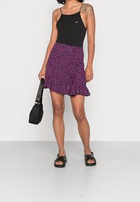 Top noir sans manches, mini jupe florale violette avec ourlet volanté, assortie à des sandales noires. Le modèle tient un sac à main noir.