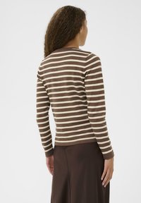 Kvinna med lockigt hår som bär en brun och beige randig långärmad topp och mörkbrun kjol, vänd bort mot en ljus bakgrund.