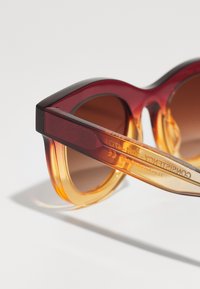 Punane ja merevaigu värvi gradientiga prillid, millel on julged ruudukujulised raamid ja pruunid läätsed. Läbipaistvad käeosad kannavad sees engraveeritud bränditeksti.