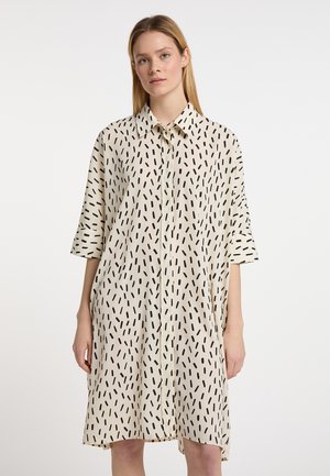 Kvinde iført en løs, knælængde beige kjole med sorte stregmønstre og trekvartærmer, stående mod en ensfarvet hvid baggrund.