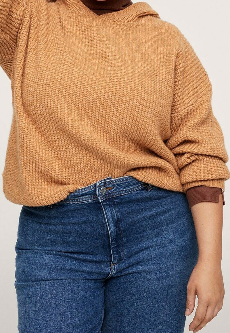 Personne portant un pull en maille marron par-dessus une chemise marron à manches longues, associé à un jean bleu taille haute, visible des épaules aux hanches.
