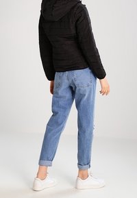 Schwarze, gesteppte Jacke mit Kapuze, kombiniert mit hellblauen, hochgekrempelten Knöchelhosen und weißen Sneakern, von hinten betrachtet.