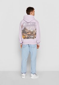 Felpa lilla con una grafica di un orso e un paesaggio forestale sul retro, abbinata a jeans azzurri chiari e sneakers bianche.
