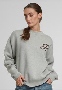 Sudadera gris de ajuste relajado, con puños acanalados y un pequeño detalle del logotipo marrón en el pecho. Textura suave y diseño minimalista.