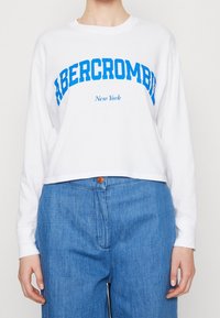 Weißes, kurz geschnittenes Langarmshirt mit blauer "ABERCROMBIE"-Schrift und "New York" darunter. Kombiniert mit blauen, hochgeschnittenen Jeans.