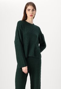 Dark green cable-knit sweater with long sleeves, round neckline, and relaxed fit, paired with matching textured pants.