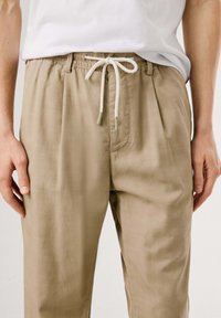 Helle beige Hose mit elastischem Bund und Kordelzug. Sie verfügt über einen plissierten Frontbereich und eine lockere Passform; hergestellt aus einem glatten, leichten Stoff.