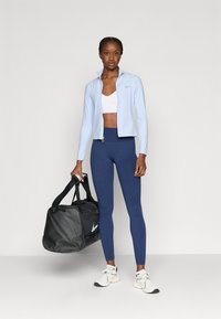 Lichtblauw jack met rits, witte sportbeha, marineblauwe legging, zwarte duffeltas en witte sneakers. Soepele stoffen en een nauwsluitend ontwerp.