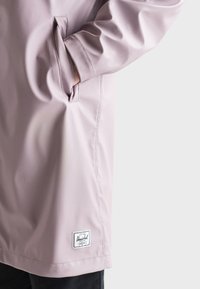 Svetloružová nepriepustná bunda s viditeľnou vreckom. Má štítok na leme a hladkú textúru. Minimalistický dizajn bez vzorov.