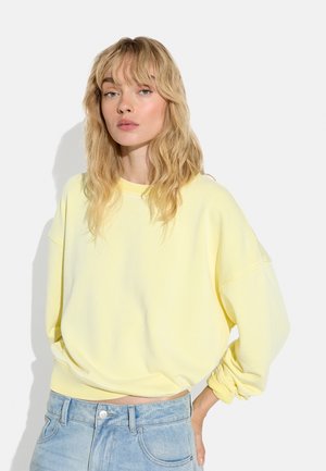 Blondynka o falowanych włosach, ubrana w luźną bladoróżową bluzę i jasnoniebieskie dżinsy, stoi na tle jednolitego jasnego tła.