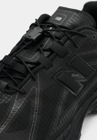 Chaussure de sport noire avec un matériau synthétique texturé, un design à lacets, une marque NLock bien visible et des accents élégants et angulaires.