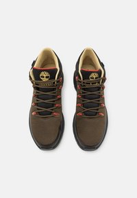 Ruskeat vaelluskengät, joissa on verkkomateriaalia ja synteettinen pinta, musta kumipohja, kultainen logo sekä punaiset yksityiskohdat nauhoissa ja kauluksessa.