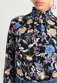 Blouse noire à motifs floraux avec un col smocké haut, mettant en valeur des fleurs et des feuilles multicolores en rose, bleu et orange sur un fond sombre.