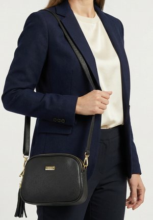 Frau trägt navyblauen Blazer und cremefarbenes Oberteil, hält eine schwarze Leder-Umhängetasche mit goldenen Beschlägen und Quasten-Detail.
