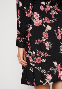 Robe noire à motifs floraux avec de vibrantes fleurs roses et blanches, manches longues avec des poignets boutonnés et un ourlet fluide.