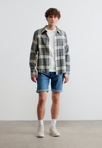 Νέος άνδρας στέκεται φορώντας γκρι καρό πουκάμισο πάνω από λευκό μπλουζάκι, τζιν σορτς με γυρισμένα μπατζάκια, λευκές κάλτσες και μπεζ αθλητικά παπούτσια μπροστά σε απλό φόντο.