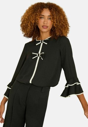 Schwarze Bluse mit weißem Piping, zwei Schleifenakzenten und Rüschensäumen. Gefertigt aus einem glatten Stoff mit lockerer Passform.