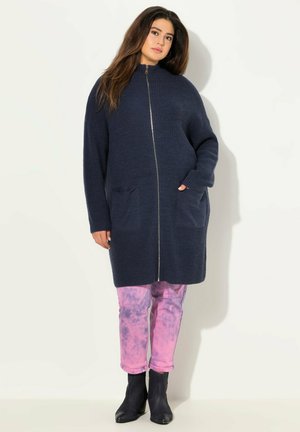 Cardigan lavorato a maglia blu con colletto alto, zip completa e due tasche frontali. Indossato sopra pantaloni rosa tie-dye e stivaletti neri alla caviglia.