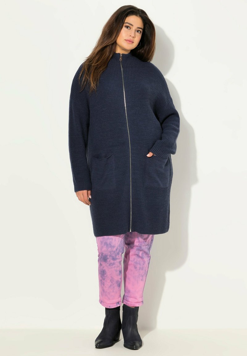 Marinblå stickad kofta med hög krage, hel dragkedja och två framfickor. Buren över rosa tie-dye byxor och svarta ankelstövlar.