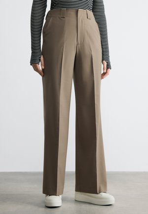 Személy, aki bézs színű, magas derekú, bő szárú nadrágot, fekete-fehér csíkos hosszú ujjú felsőt és fehér platform sportcipőt visel, beltéren áll.