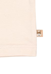 Helles beige Baumwoll-T-Shirt mit einem gesäumten Rand und einem kleinen Stoffetikett, das ein lächelndes Tiergesicht zeigt.