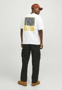 T-shirt bianco con una grafica nera e gialla sulla schiena, abbinato a pantaloni cargo neri e stivali marroni. Stile streetwear casual.