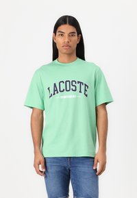 Mėtų žalios spalvos medvilnės marškinėliai su tamsiai mėlynu bold rašmenimis "LACOSTE SPORTSWEAR" logotipu, trumparankoviai ir apykaklė.