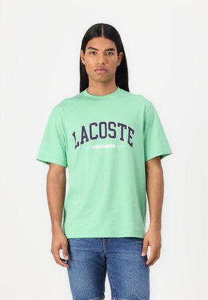 Mint green cotton t-shirt featuring a navy blue "LACOSTE SPORTSWEAR" logo in bold print, with short sleeves and a crew neckline.