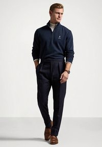 Camisola de malha com fecho éclair azul-marinho, camisola de gola alta cinza clara, calças com padrão risca-de-giz escuras e ajustadas, mocassins castanhos e um relógio com correia castanha.