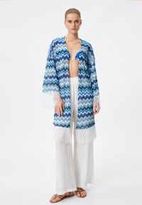 Blauw en wit gestreepte kimono-cover-up met zigzagpatroon, franjes aan de onderkant, gecombineerd met witte wijde broek en bruine sandalen.