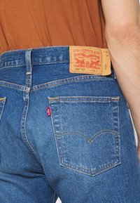 Blå jean byxor i denim med en brun läderlapp som visar "Levi Strauss & Co." och stilnummer. Har en röd etikett och gula sömmar på fickorna.