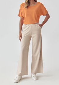 Volná oranžová tričko s širokým výstřihem, doplněná béžovými kalhotami s širokými nohavicemi. Tkanina se zdá být měkká, s hladkým texturem a jemnými švy.