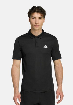 Mann trägt ein schwarzes Adidas-Poloshirt mit weißem Logo, steht vor einem schlichten hellen Hintergrund, blickt nach vorne mit neutralem Gesichtsausdruck.