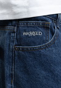 Mørkeblå denimjeans med teksturert overflate. Har "WASTED" brodert på baklommen med hvit tråd, og kobbernagler.