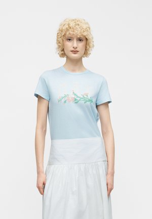 Rövid, göndör szőke hajú személy világoskék, virágmintás pólóban és fehér szoknyában, egyszínű háttér előtt.