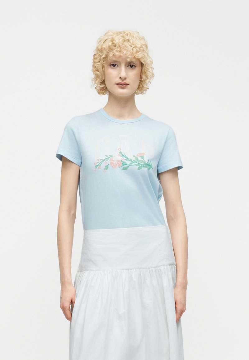 Άτομο με κοντά σγουρά ξανθά μαλλιά που φοράει ένα γαλάζιο μπλουζάκι με σχέδιο λουλουδιών και μια λευκή φούστα, μπροστά από μια απλή φόντο.