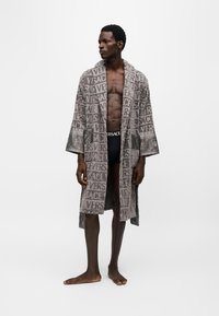 Мъж, стоящ бос, облечен в сив халат Versace с лого и черно бельо Versace на бял фон.