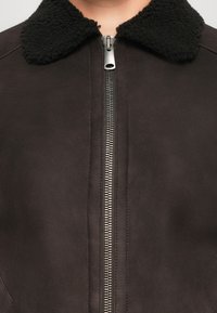 Läheltä kuvattu tumma ruskea takki, jossa on musta pörröinen kaulus ja hopeinen metallizipper, vetoketju joka on vedetty kaulaan saakka.