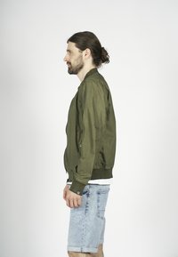Giacca bomber verde oliva con polsini e orlo a coste, chiusura con zip e tasche laterali, abbinata a pantaloni corti in denim chiaro.