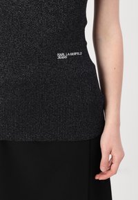 Crni rebrasti top s metalnim nitima, s prilagođenim siluetom i "KARL LAGERFELD JEANS" logotipom u bijeloj boji. Glatka tekstura, minimalistički dizajn.