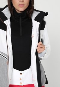 Femme portant un haut noir zippé sous une veste de ski blanche et grise, tenant la veste ouverte, avec un pantalon rouge visible en bas.