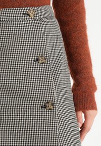 Jupe pied-de-poule noir et blanc avec quatre boutons dorés, taille haute, et pull en laine texturé de couleur rouille. Vue en gros plan.