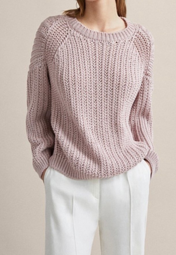 Femme portant un pull en maille épaisse rose clair à col rond et un pantalon blanc taille haute, mains dans les poches.