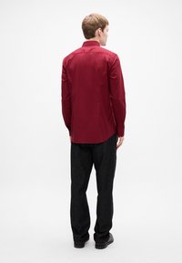 Uomo in piedi di spalle, indossa una camicia a maniche lunghe bordeaux, pantaloni scuri e scarpe nere, su uno sfondo bianco uniforme.