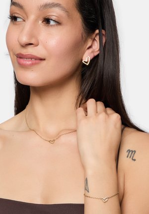 Goldschmuckset mit herzförmigen Ohrringen, einer passenden Halskette mit einem kleinen Herzanhänger und einem zarten Armband, alles auf einem hautfarbenen Hintergrund.